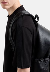 Zaino in pelle nera con finitura testurizzata, spallacci regolabili e chiusura arricciata, indossato sopra una t-shirt nera con marchio in rilievo.