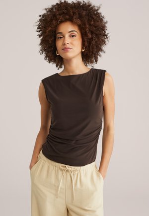 Barna ujjatlan felső sima anyagból, oldalán ráncolt részletekkel, világos bézs nadrággal kombinálva, amelyen húzózsinóros derékrész található.