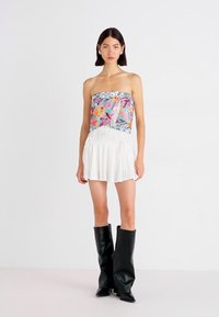 Strapless top met bloemenpatroon, versierd met kralenfranje, gecombineerd met een witte rok; model draagt hoge zwarte leren laarzen. Eenvoudige achtergrond.