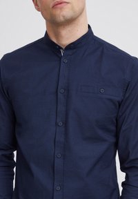 Camisa azul marino de botones con cuello de banda, de mangas largas y un bolsillo en el pecho. Hecha de un tejido texturizado con botones a juego.