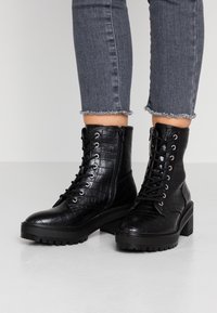 Zwarte enkellaarsjes van kunstleer met krokodillenstructuur, ronde neus, veters aan de voorkant en een chunky hak. Versleten spijkerbroek met rafelige zoom.