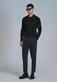 Polo noir à manches longues côtelé associé à un pantalon plissé gris foncé. Une ceinture noire et des chaussures à lacets complètent la tenue. Des lunettes de soleil sont portées.