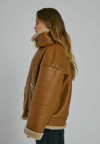 Vrouw met lang haar, gekleed in een bruin leren jack met beige schapenwollen voering, staande in profiel tegen een effen grijze achtergrond.