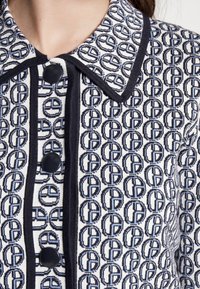 Zbliżenie wzorzystej koszuli z tkaniny z czarnymi okrągłymi logo, czarnymi lamówkami oraz dużymi czarnymi guzikami na przodzie i kołnierzu.