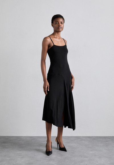 Rochie neagră fără mâneci, cu corset ajustat și fustă evazată. Dispune de o deschidere laterală de la genunchi și are un material neted. Este asortată cu pantofi cu toc negri.