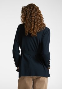 Persona con cabello rizado que lleva una chaqueta oscura de navy con cinturón, puños abotonados y pantalones marrón claro, de espaldas frente a un fondo plano.