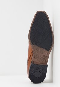 Chaussure en cuir marron avec une semelle en caoutchouc noir texturé. Présente un dessus lisse et une zone rembourrée à l'arrière pour plus de confort.