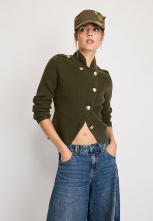Femme portant un pull boutonné vert olive, un jean large, une chemise à carreaux nouée à la taille, des ballerines beiges et une casquette verte olive assortie sur fond blanc.