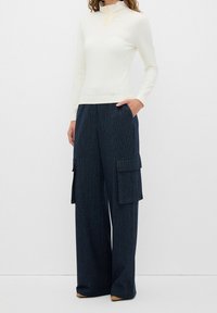 Biały top z długim rękawem, wysokim kołnierzem i małym detalem w postaci guzika, zestawiony z granatowymi szerokimi spodniami typu cargo w cienkie prążki, z bocznymi kieszeniami.