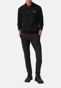 Mann, der einen schwarzen Pullover mit strukturiertem Strickmuster über einem roten Hemd, dunklen grauen Hosen und schwarzen Schnürstiefeln trägt, steht mit den Händen in den Taschen.