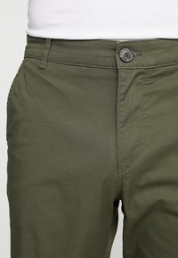 Oliwkowe bawełniane spodnie z zapięciem na guzik, standardowym paskiem, przednimi kieszeniami i gładką teksturą z subtelnym wzorem w jodełkę.