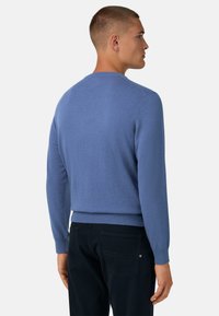 Mėlynas megztinis su ilgomis rankovėmis ir apvalia apykakle. Minkšta tekstūra, prigludęs siluetas ir raukšlėti rankogaliai. Derinama su tamsiomis kelnėmis.