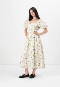 Keltainen kukallinen mekko, jossa on puffihihat, istuva vartalo ja virtaava hame. Sisältää sinisiä kukkia ja teksturoitua kangasta. Mustat kengät jalassa.