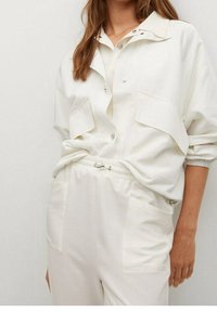 Witte, losse blouse met een puntige kraag, knoopsluiting en twee borstzakken. Gecombineerd met bijpassende elastische broek met zijzakken.