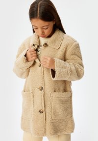 Beige teddy-frakke med krave, træknapper og store frontlommer. Teksturmateriale, blødt og med en afslappet pasform samt lange ærmer.