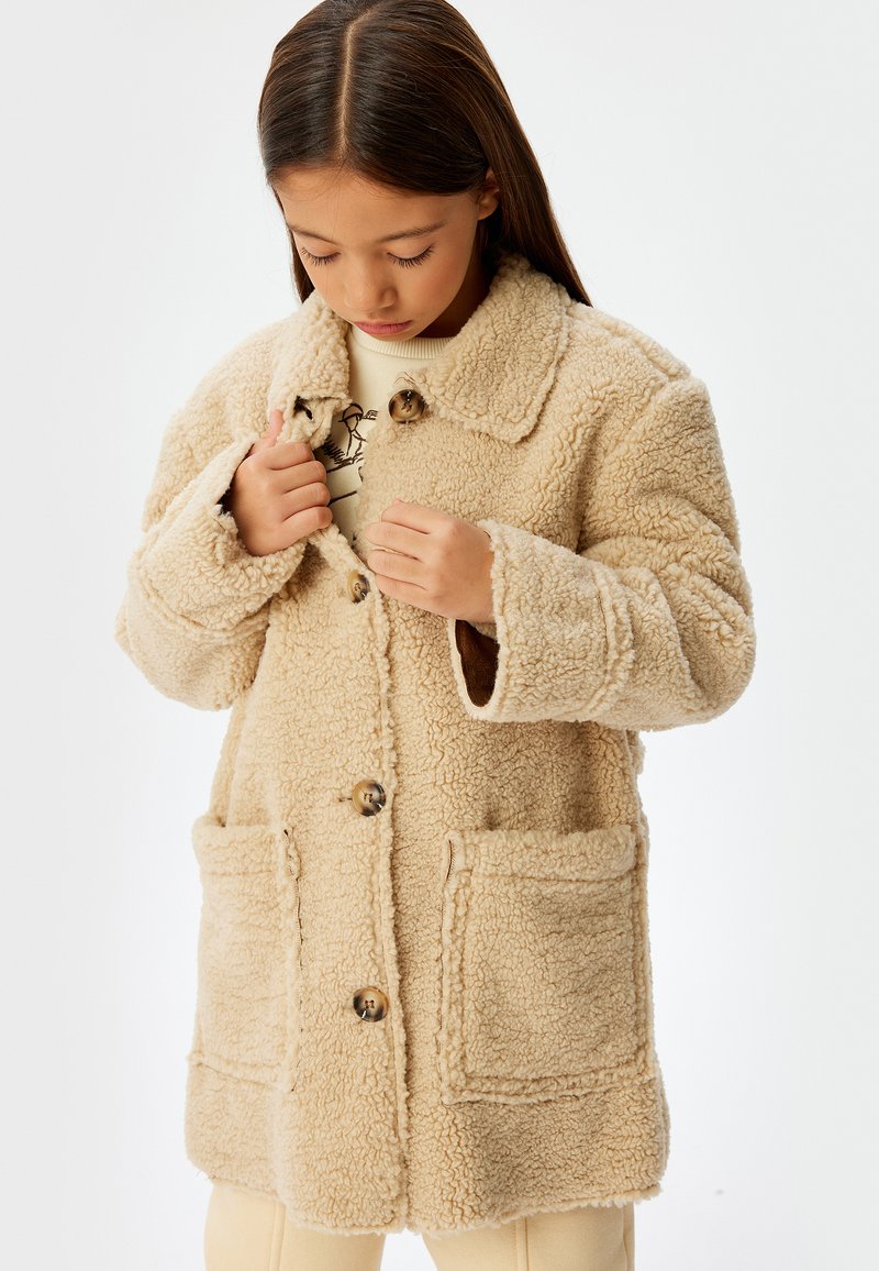 Beige teddy-frakke med krave, træknapper og store frontlommer. Teksturmateriale, blødt og med en afslappet pasform samt lange ærmer.