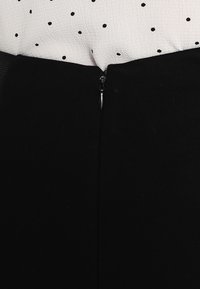 Zwarte rok met een gladde textuur, ritssluiting aan de achterkant en een aansluitend ontwerp, gedeeltelijk zichtbaar met een blouse met stippen bovenaan.