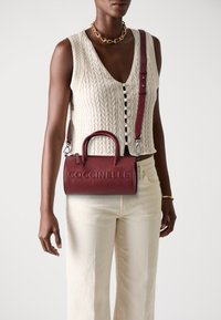 Red leather handbag with a textured finish, rounded handles, and a detachable strap, featuring embossed "Coccinelle" logo on the front.