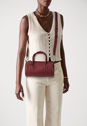 Red leather handbag with a textured finish, rounded handles, and a detachable strap, featuring embossed "Coccinelle" logo on the front.