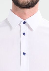 Camisa de cuello blanco con botones azules, tejido suave y diseño clásico. La vista en primer plano resalta los detalles de los botones y la textura.