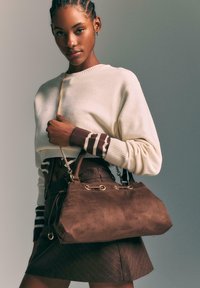 Sac à main en suède brun avec des accessoires en or, doté de deux poignées et d'une bandoulière en chaîne, accompagné d'un léger pull et d'une jupe à rayures brunes.