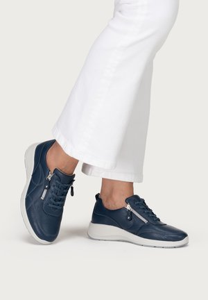 Füße in dunkelblauen Leder-Sneakern mit Schnürsenkeln und seitlichen Reißverschlüssen, kombiniert mit weißen gekürzten Hosen.