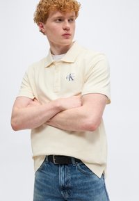 Νέος άνδρας με σγουρά κόκκινα μαλλιά, ντυμένος με μια μπεζ πόλο μπλούζα Calvin Klein και μπλε τζιν, στέκεται με σταυρωμένα τα χέρια, κοιτάζοντας προς τα πλάγια.