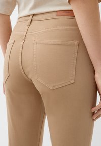 Beżowe dżinsy elastyczne o dopasowanym kroju, z dwoma kieszeniami z tyłu i naszywką z logo w pasie. Gładka tekstura i minimalne detale przeszyć.