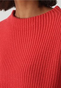 Nahaufnahme einer Person, die einen leuchtend roten, gerippten Pullover mit Rundhalsausschnitt trägt und kurzhaarsichtig ist.