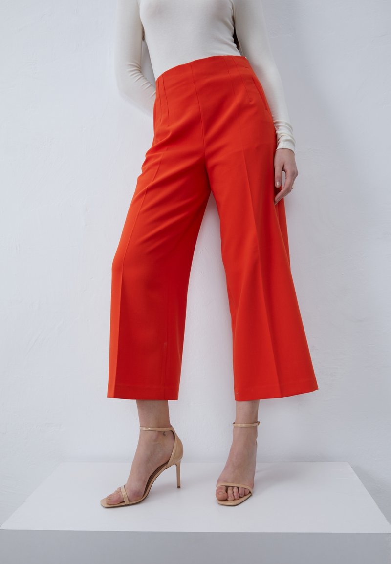 Personne portant un pantalon taille haute, à jambes larges, de couleur orange vif, avec des sandales à talons beiges, posant devant un mur blanc uni.