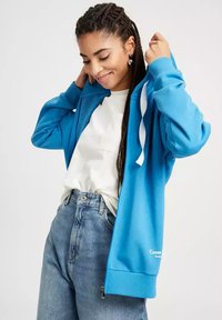 Niebieska bluza z kapturem z zamkiem z przodu, białe sznurki, ściągacze przy rękawach oraz detal z logo u dołu, zestawiona z jasnymi jeansami o wysokim stanie.