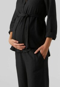 Crna platnena majica za trudnice s gumbima, pojasom i manžetama na gumb, uparena s odgovarajućim crnim hlačama s bočnim džepovima.
