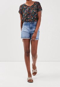 Mujer con una blusa de flores de manga corta, shorts de mezclilla de tiro alto de color azul claro y sandalias beige con tiras entrecruzadas caminando hacia adelante.