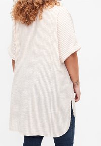 Beige und weiße gestreifte übergroße Bluse mit kurzen Ärmeln, abgerundetem Saum und seitlichen Schlitzen, aus einem leichten Stoff gefertigt.