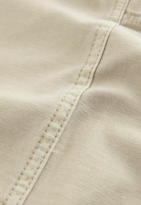 Beige stof med en glat tekstur, der har synlige sømme langs kanten, der viser et tæt billede af en sømdetalje.