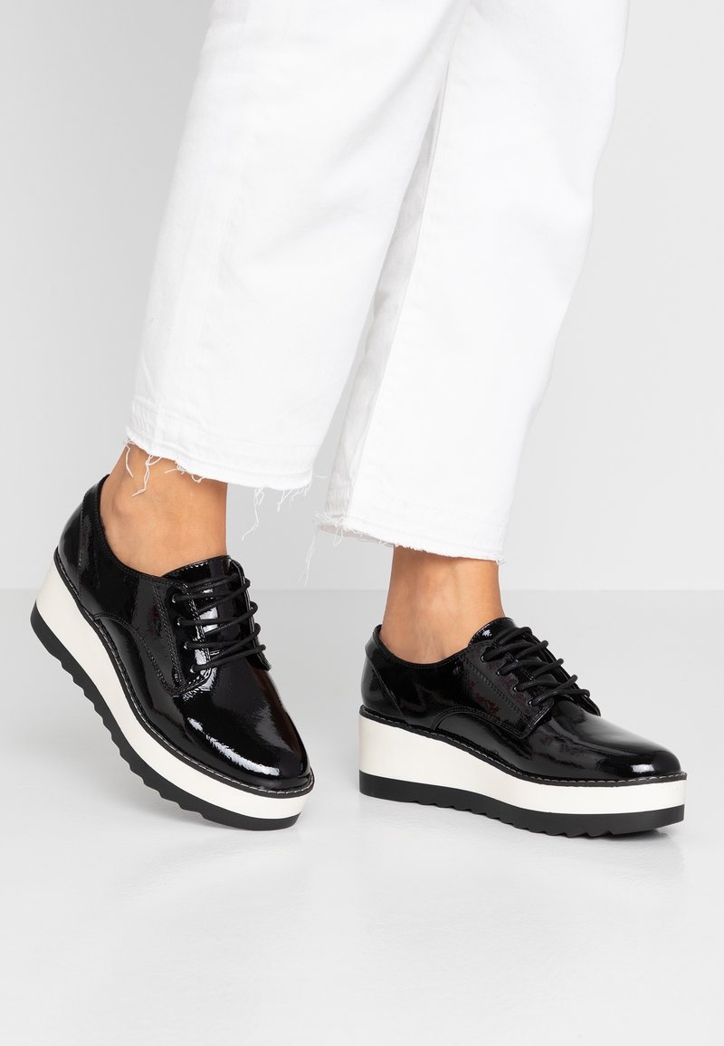 Chaussures à plateforme en cuir verni noir avec design à lacets ; dotées d'une semelle intermédiaire blanche et d'une semelle extérieure noire texturée pour une meilleure adhérence.