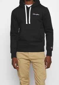 Black cotton hoodie with a front kangaroo pocket, white drawstrings, and a small logo on the chest. Long sleeves with ribbed cuffs.