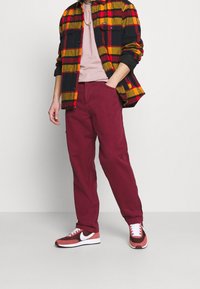 Mann in kastanienbraunen Hosen, mit einer rot-gelben karierten Jacke über einem pinken Hemd und roten, weißen sowie braunen Sneakers, der mit einer Hand in der Tasche steht.