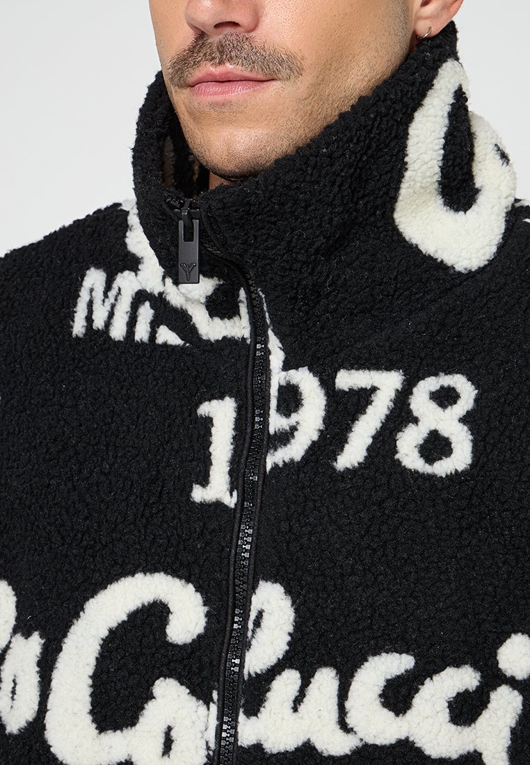 Schwarze Fleecejacke mit weißem Text, hohem Kragen mit Reißverschluss und flauschiger Textur. Besondere Merkmale sind das Jahr "1978" und Markenelemente.