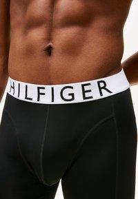 Ropa interior negra elástica con una cinturilla blanca que presenta "HILFIGER" en letras negritas negras. Textura suave, diseño ajustado y sin patrones visibles.