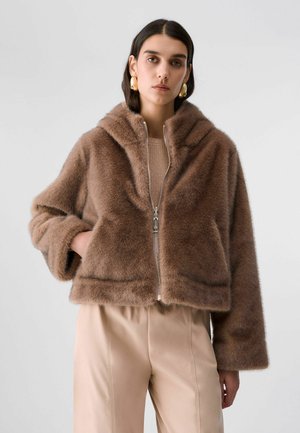 Donna che indossa una giacca marrone pelosa con zip e tasche laterali, pantaloni beige e grandi orecchini a cerchio dorati, in piedi davanti a uno sfondo semplice.