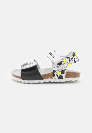 Sandale pour enfant avec sangle avant noire, sangle de cheville blanche avec imprimés de personnages de dessin animé, semelle en liège et fermetures velcro blanches.