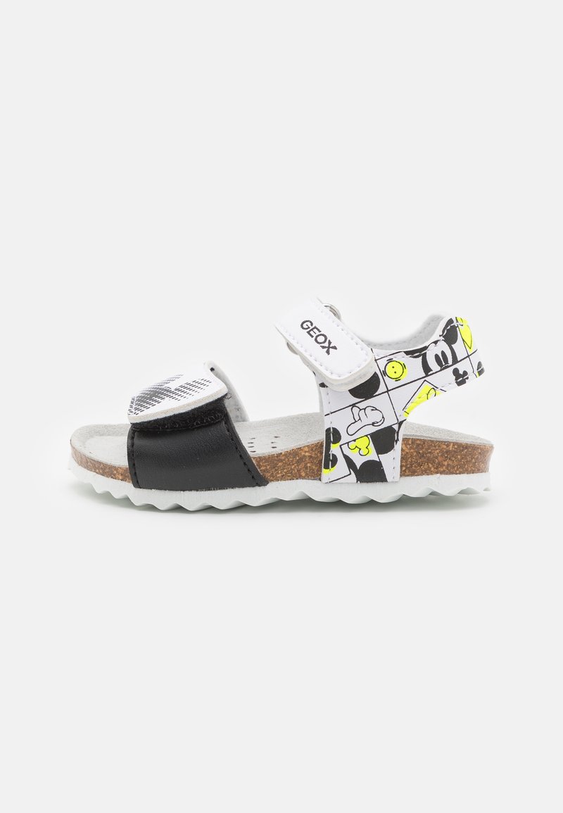 Sandale pour enfant avec sangle avant noire, sangle de cheville blanche avec imprimés de personnages de dessin animé, semelle en liège et fermetures velcro blanches.