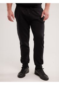 Mees, kes kannab musti jooksepükse elastsete mansettidega ja musti tossusid, seisab lihtsa heleda tausta ees.