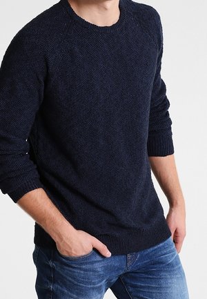 Mann trägt einen dunkelblauen strukturierten Strickpullover mit gerippten Bündchen und Saum, kombiniert mit mittelblauen Denim-Jeans, Hände in den Taschen.