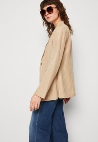 Blazer beige a righe con un solo bottone, abbinato a jeans in denim a gamba larga. Il tessuto ha una texture liscia e una vestibilità rilassata.