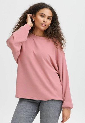 Pulover oversized roz deschis, cu mâneci lungi, ușor volă, și guler rotund, asortat cu blugi gri. Textură moale și netedă a materialului.