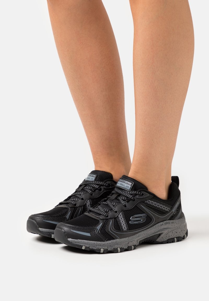 Jambes inférieures portant des chaussures de course sur sentier Skechers noires et grises avec des semelles à motifs sur fond blanc.