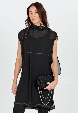 Donna con capelli castani indossa un abito nero senza maniche con scollo trasparente, tiene in mano una pochette nera con borchie e catene d'argento.