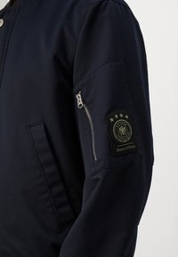 Rękaw marynarskiej kurtki z kieszenią na suwak i czarną naszywką z czterema gwiazdkami, emblematem orła oraz napisem „Marc O'Polo”.